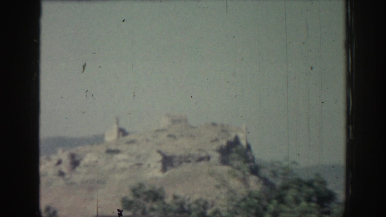 1962 Rhodes frame showing distant hilltop ruins above scrub vegetation, with grain and vertical wear marks in the film.