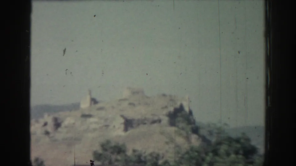 1962 Rhodes frame showing distant hilltop ruins above scrub vegetation, with grain and vertical wear marks in the film.
