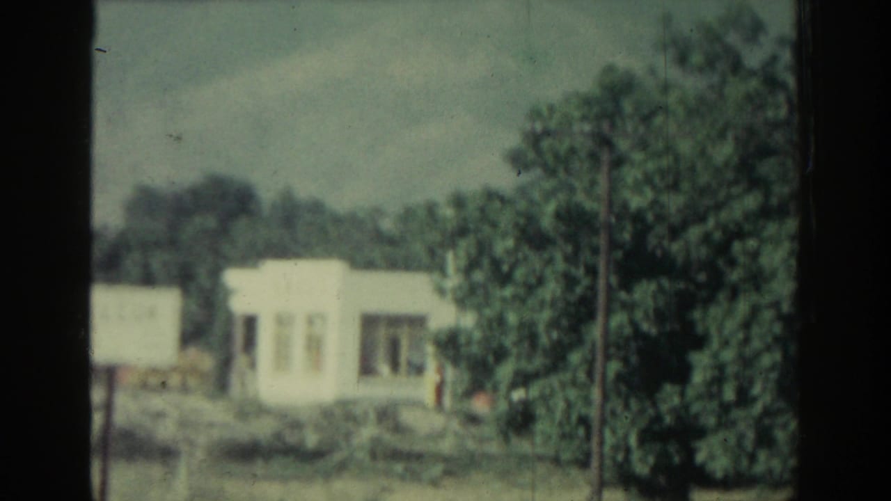 White roadside building, trees and low landscape seen through a car window or film frame edge.