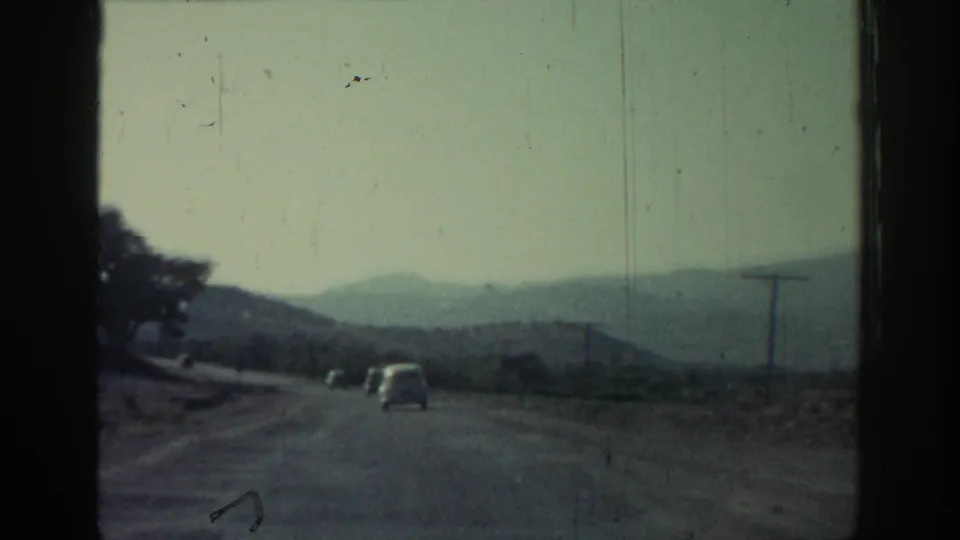 1962 8mm road-trip frame showing cars ahead on a rural road with hazy hills and open roadside landscape.