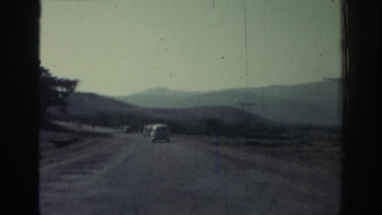 1962 8mm view from a moving vehicle on a mountain road in Rhodes, Greece, with a small car ahead and hazy hills beyond.