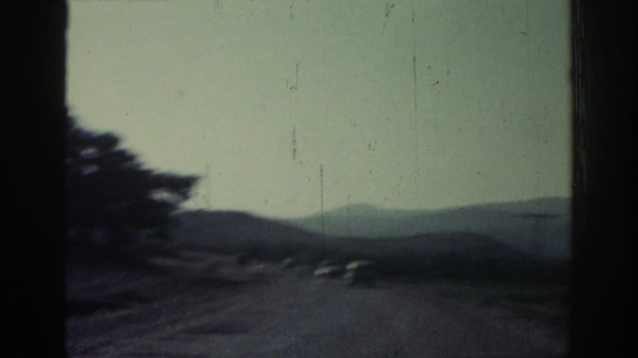Vehicle-perspective 8mm frame from Rhodes, Greece, showing a curving mountain road with trees, hills, and distant cars.