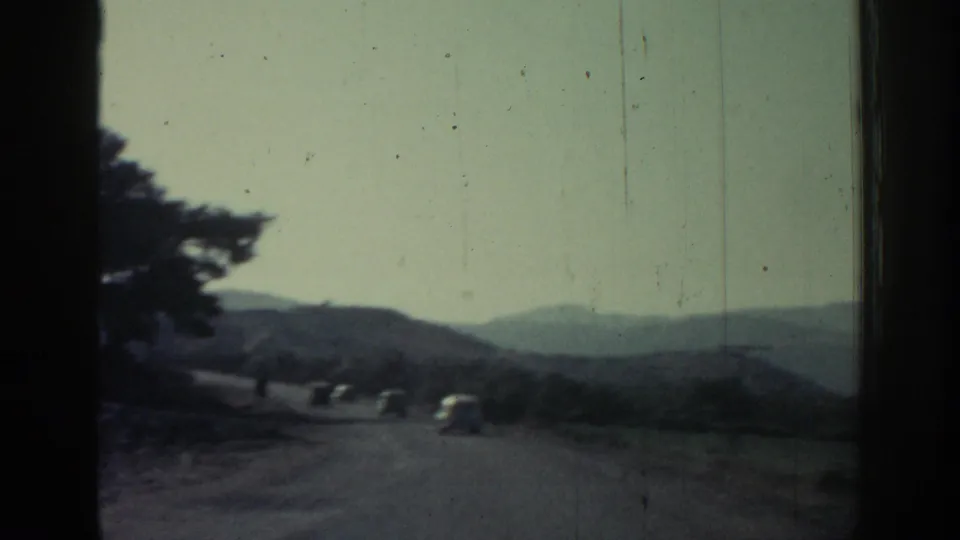 1962 Rhodes road-travel frame with several small cars on a mountain road beneath a pale sky and distant hills.