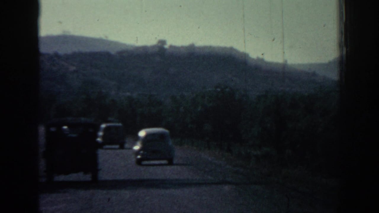 Classic cars drive along a road bordered by hills and vegetation.