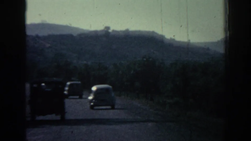 1962 Rhodes home-movie frame showing cars on a hilly road through green vegetation.