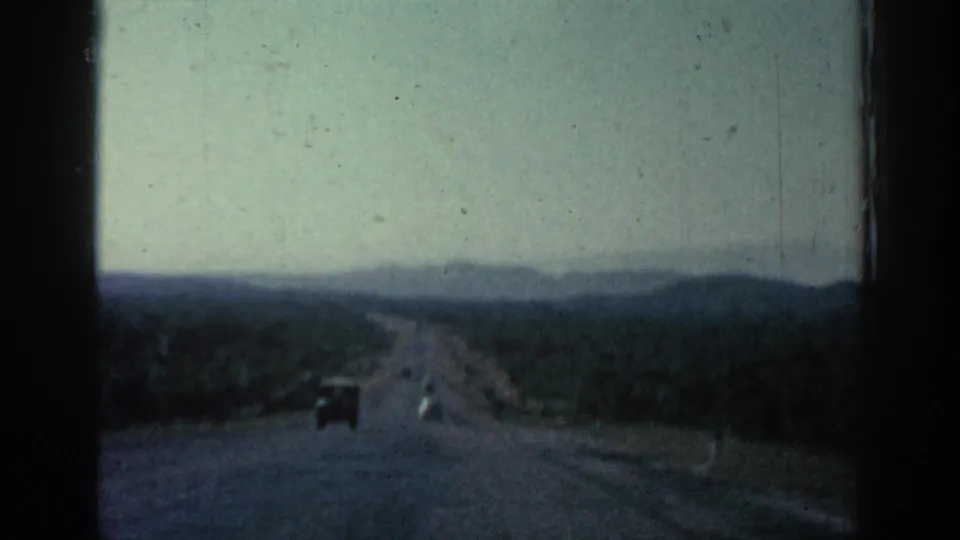 Rhodes road scene from a moving vehicle, with one car ahead and hills beyond.