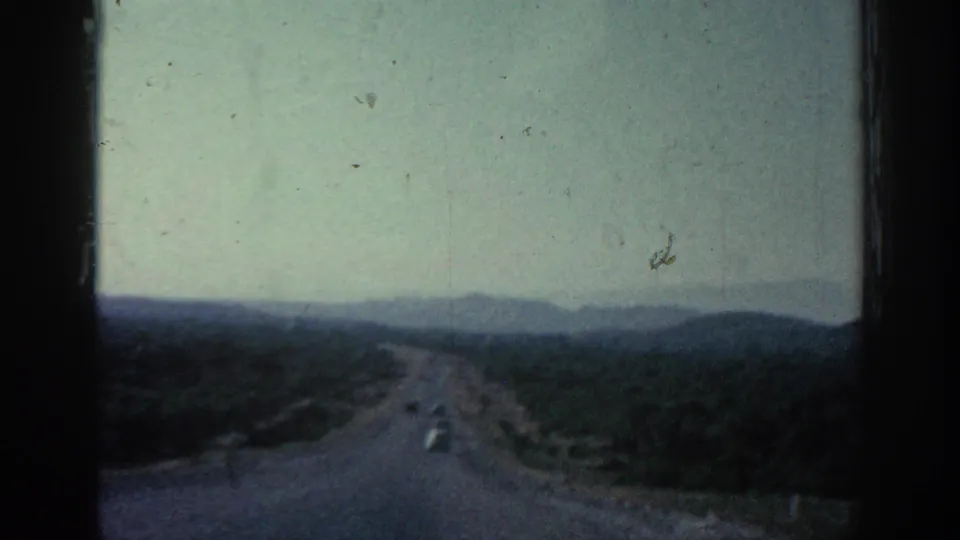1962 Rhodes travel footage frame showing a winding hillside road and distant car.