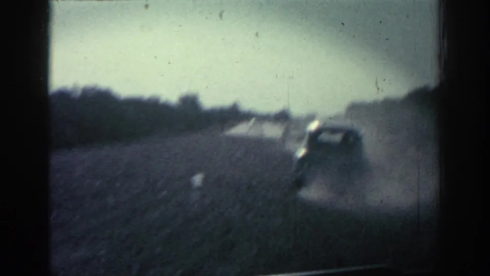 1962 Rhodes road-travel frame with a car ahead on a gravel surface, dust rising across the right side of the view.