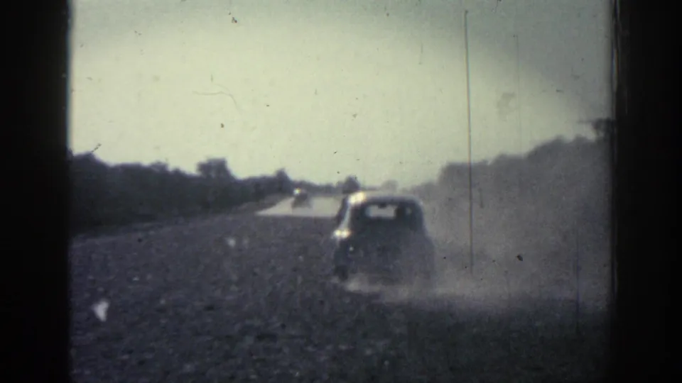 1962 Rhodes 8mm still with a car ahead on a dusty rural road, distant landscape, and visible film wear.