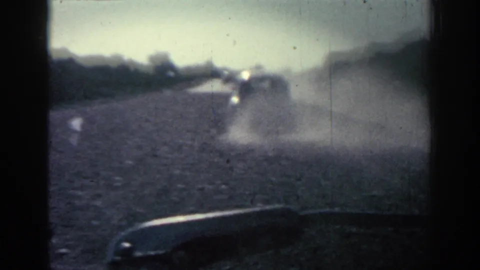 1962 Rhodes road footage frame showing a car through dust, with part of the filming vehicle or window edge at the bottom.