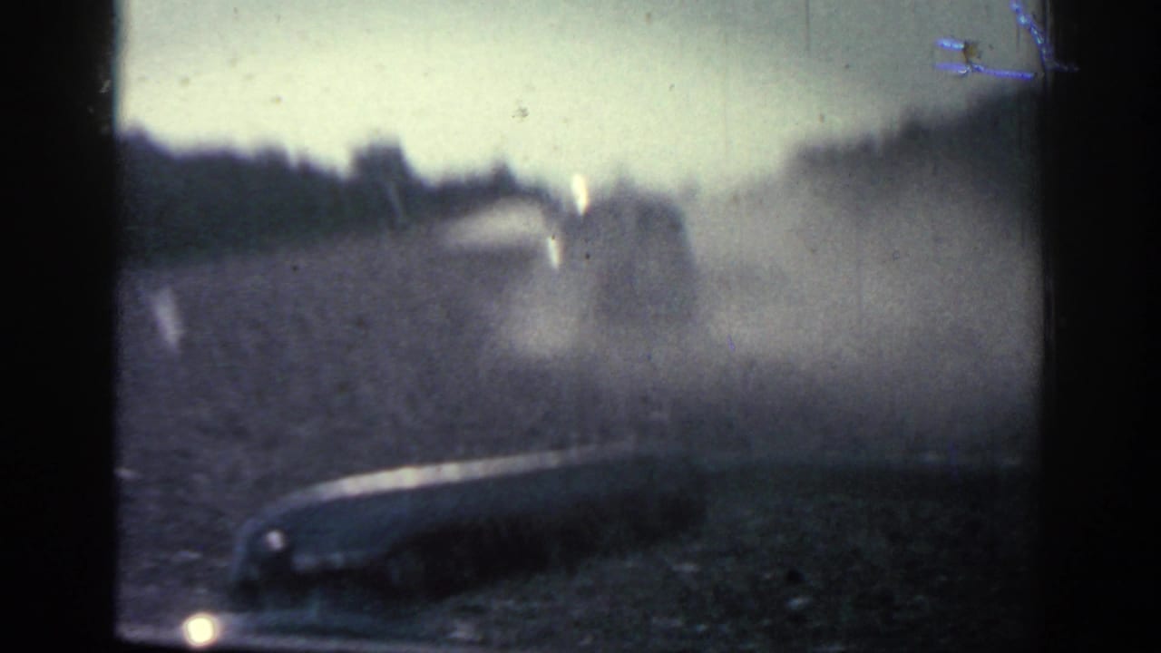 1962 Rhodes home-movie frame showing a dusty road through a windshield or open vehicle view, with a vehicle ahead and trees along the road.