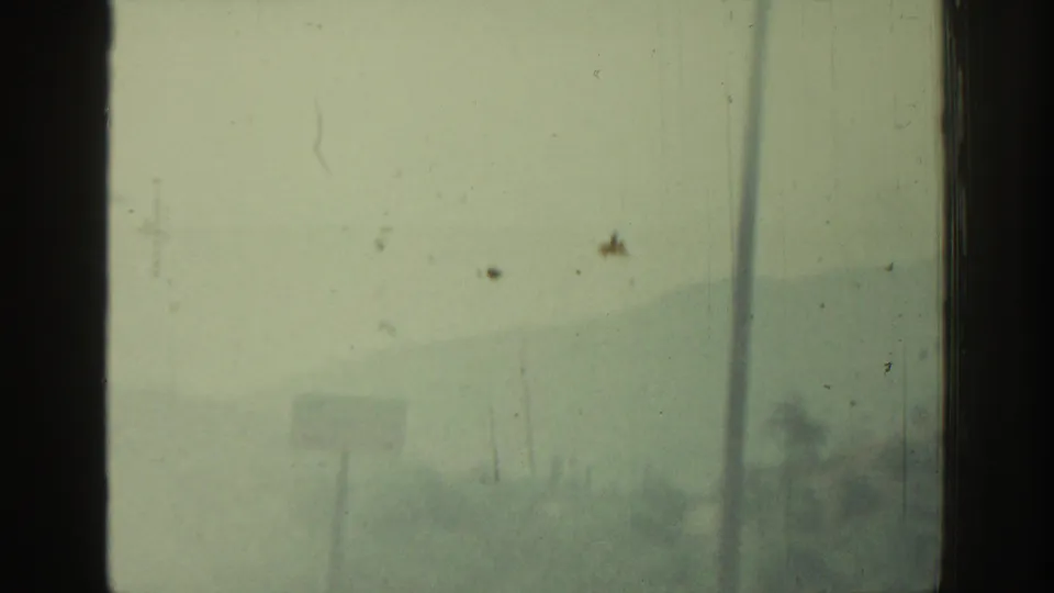 1962 Rhodes home-movie frame showing a pale, dusty landscape through dark vehicle-window divisions and scratched 8mm film.