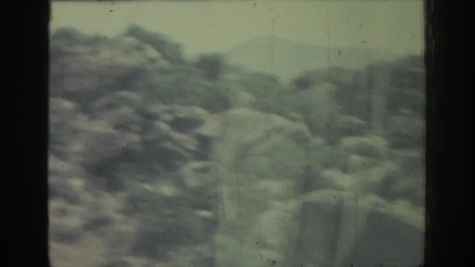 Rock-strewn hillside and distant terrain in Rhodes, Greece, filmed softly from a moving car on 8mm in 1962.