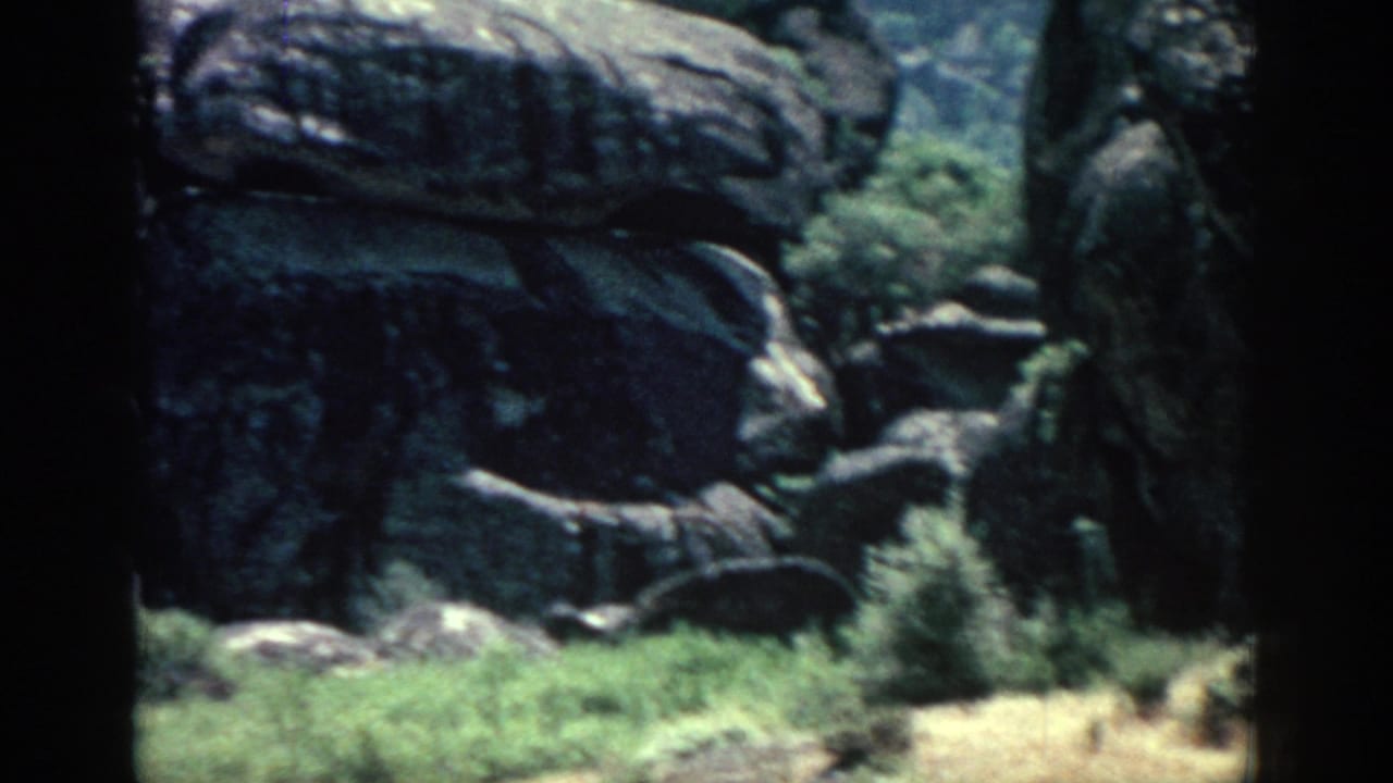 1962 Rhodes home-movie frame showing weathered boulders, shrubs, dry ground, and dark 8mm frame edges.