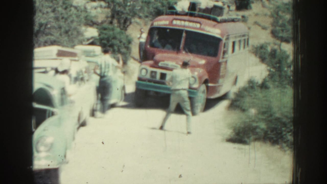 Red bus facing a narrow road as a man directs traffic beside parked cars.