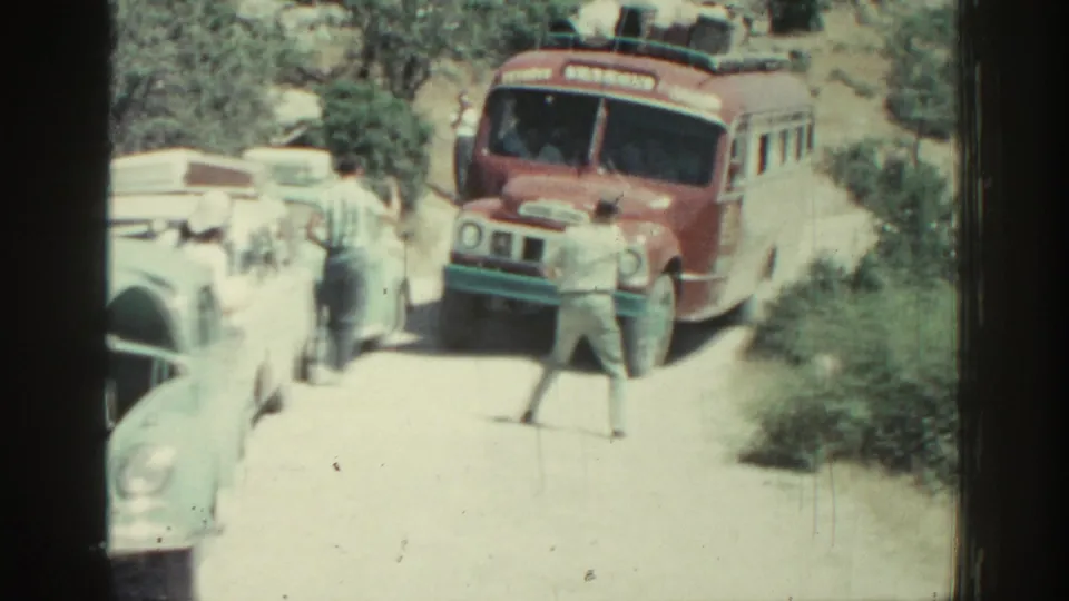 1962 Rhodes, Greece home-movie frame showing a red bus approaching while a man guides traffic on a dusty narrow road.