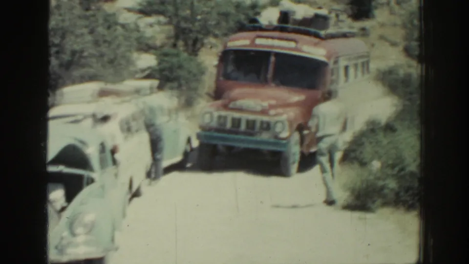 A red bus noses forward on a narrow Rhodes road in 1962, with cars close by and a man positioned to guide it through.