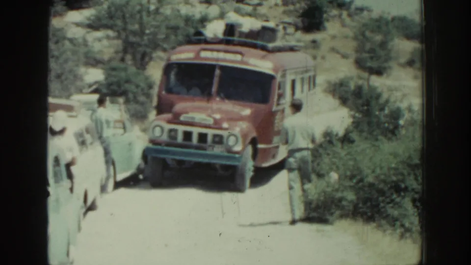 The red bus is closer to the camera, passing light-colored cars as people stand along the narrow Rhodes roadside.