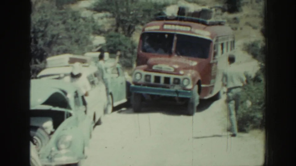 A red bus advances through tight traffic on a Rhodes road in 1962, with a man nearby and pale cars lining the route.
