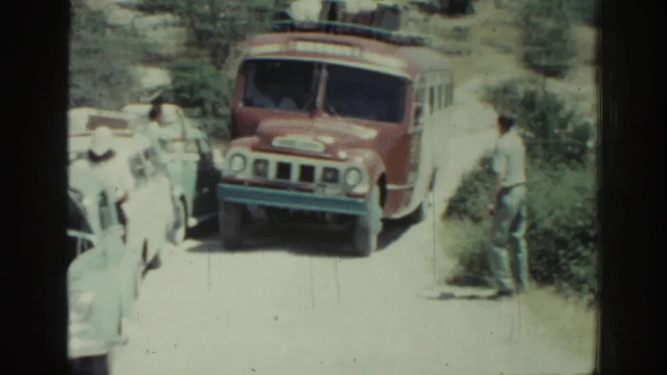 Red-and-white bus in Rhodes, Greece, with two men near the driver area and pale cars crowding the roadside.