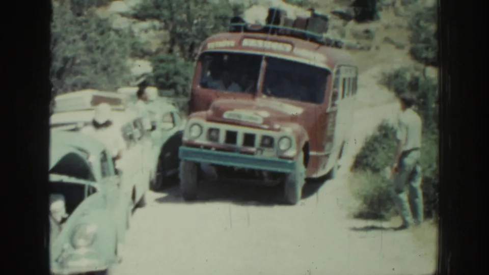1962 Rhodes frame with the bus angled through a narrow gap beside classic cars and a man watching from the roadside.