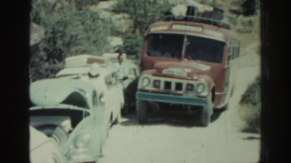 Red-and-white bus and pale classic cars share a constricted Rhodes roadside in a 1962 home-movie frame.
