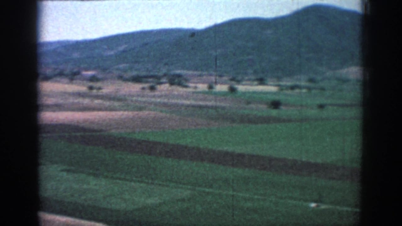 1962 Rhodes-area countryside appears from a moving vehicle, with fields, hills, and dark window framing.