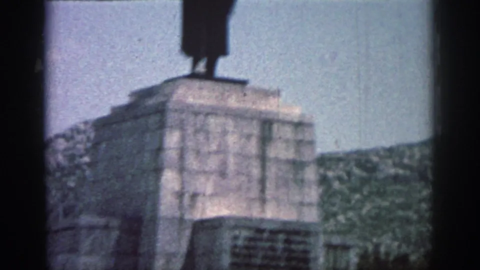 Visitor-shot 8mm view of a tall grey monument in Rhodes, with a dark statue silhouette above the base.