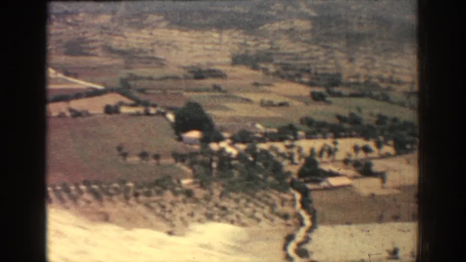 1962 Rhodes travel-film frame looking across cultivated fields, paths, small structures, and hills beyond.