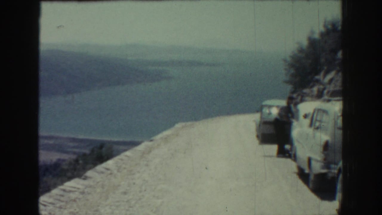 Cliffside road overlooking water and hills with parked vehicles and a person near a van.