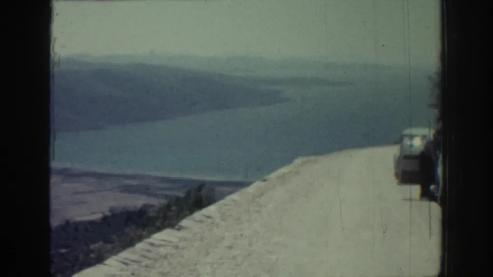 1962 Rhodes view from a roadside overlook, with parked vehicles on the right and a broad bay or water body below distant hills.
