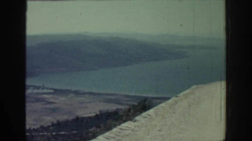 1962 Rhodes scenic frame looking from a high road or parapet toward water, low shoreline, and distant hills.