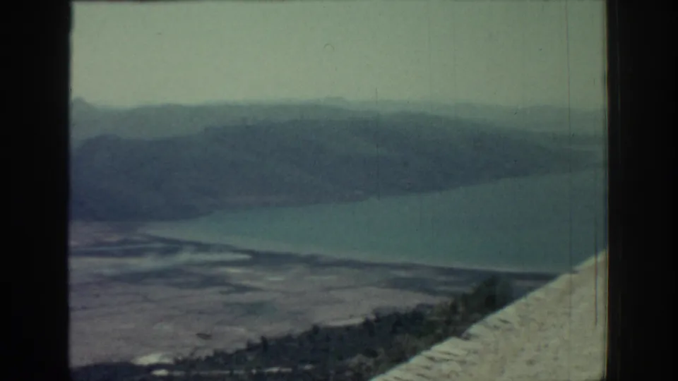 1962 Rhodes home-movie frame of water, distant hills, shoreline, and a pale cliffside or road edge at the right.