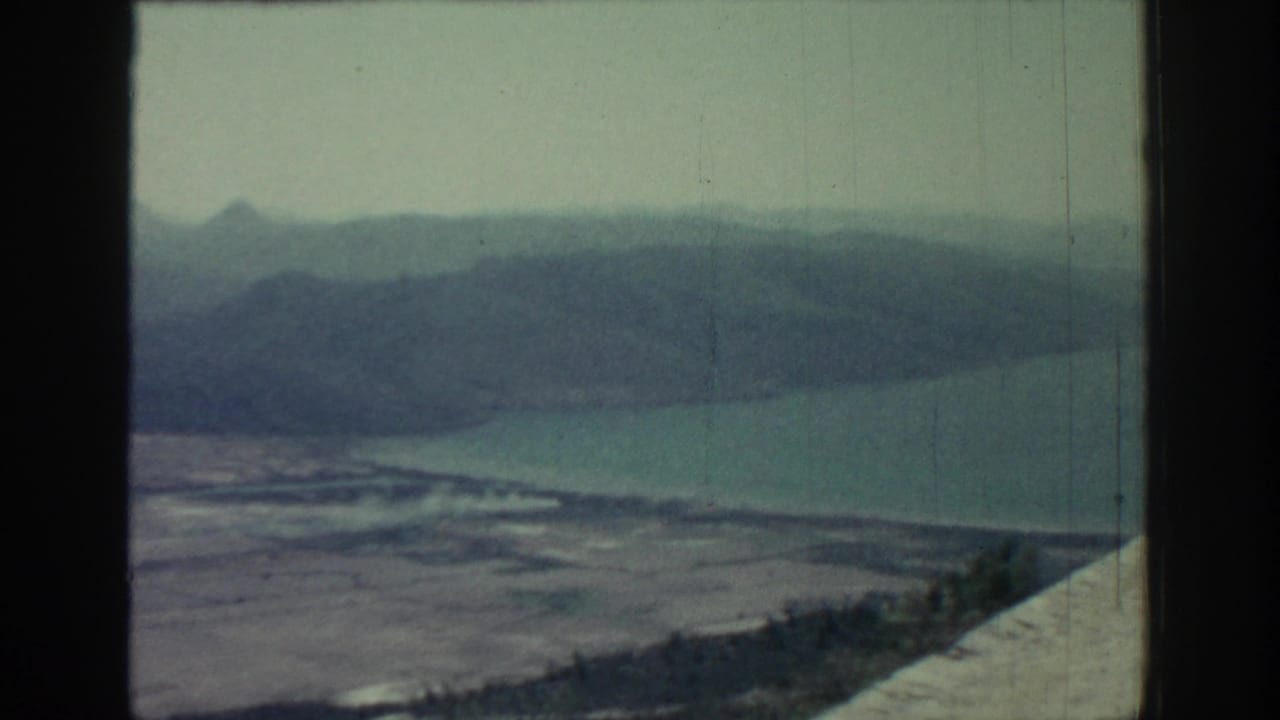 Elevated coastal view with sea, distant mountains, pale shoreline, cloudy sky, and dark film borders.