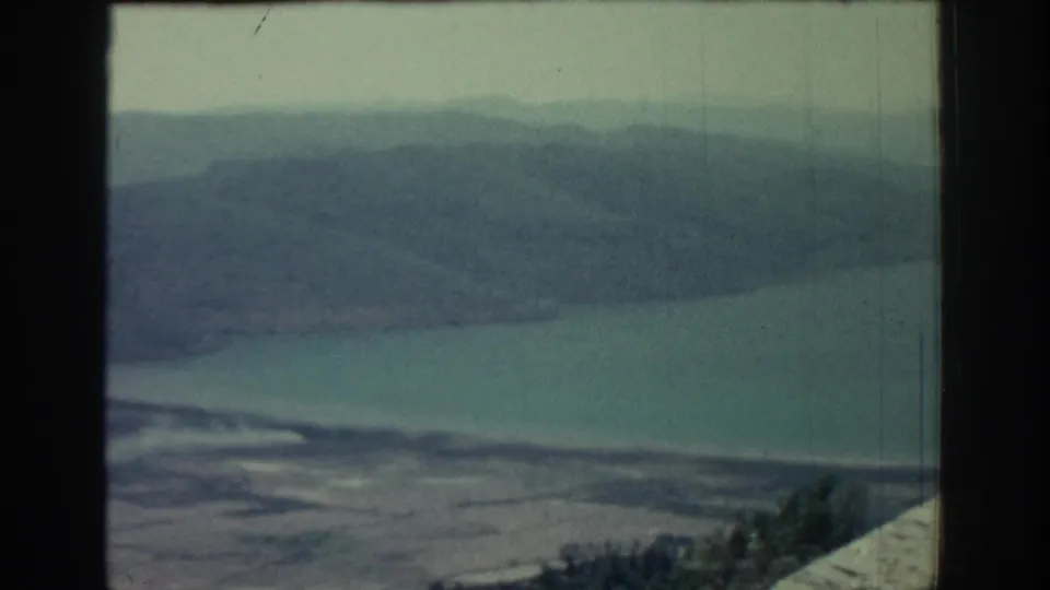 Elevated 1962 Rhodes coastal view with distant hills, calm sea, pale beach or shore, cloudy light, and dark projector-frame edges.