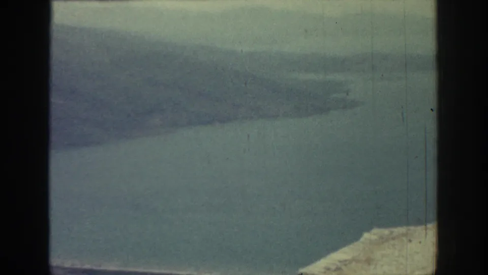 1962 Rhodes frame with muted sea, distant hazy mountain ridge, cloudy sky, pale shoreline at lower right, and visible 8mm wear marks.