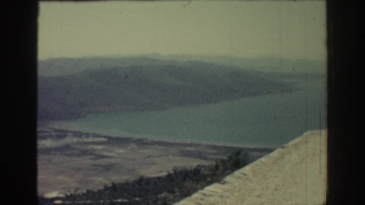 1962 Rhodes frame with a blue-green coastal inlet, layered hills, sparse trees, and pale rock at the right edge.