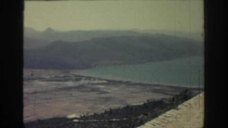 1962 Rhodes home-movie view of water below distant mountains, with open shore terrain and a strip of dark vegetation.