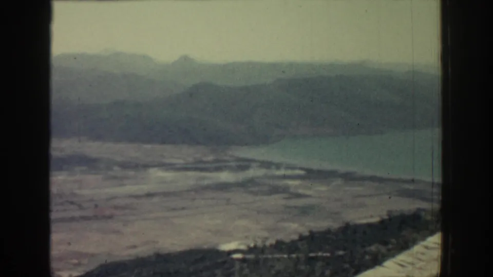 1962 Rhodes frame showing layered mountain silhouettes, calm coastal water, rocky ground, and sparse vegetation under haze.