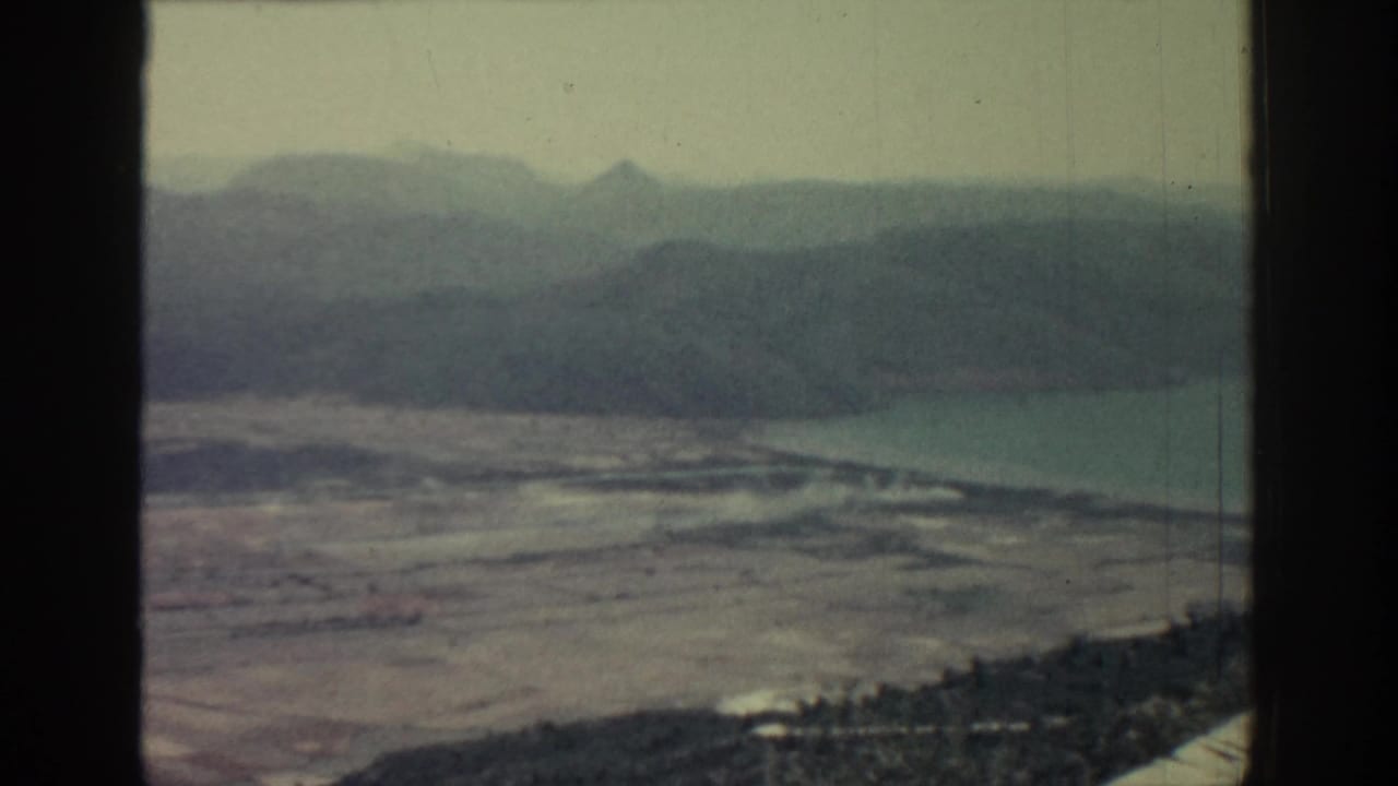 1962 Rhodes home-movie frame showing a soft, hazy coastal landscape with fields, water and layered mountains beyond.