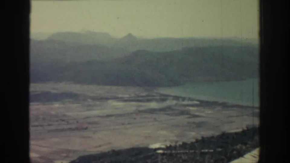 1962 Rhodes home-movie frame showing a soft, hazy coastal landscape with fields, water and layered mountains beyond.