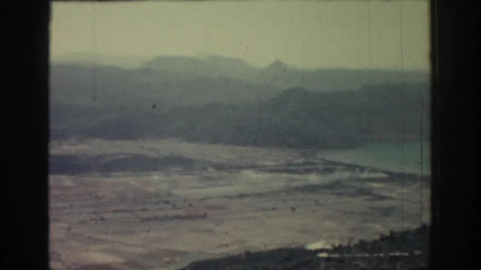 Wide 1962 Rhodes landscape frame with muted 8mm color, pale beach or fields, water and hazy mountains in the distance.