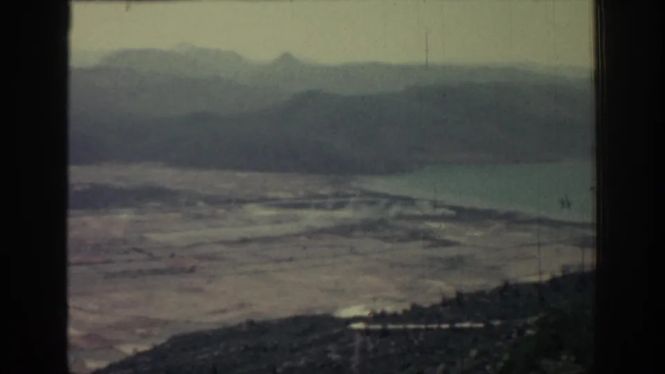 Final 1962 Rhodes frame showing a distant coastal plain, water, dark foreground hillside and layered mountains in soft home-movie color.