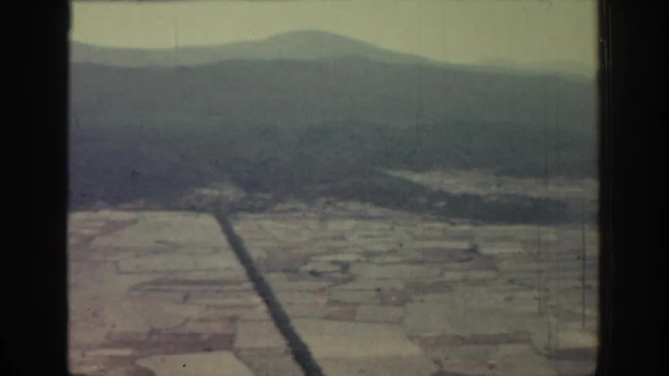 1962 Rhodes aerial frame with a dark road or channel crossing tan fields below hazy mountain slopes.