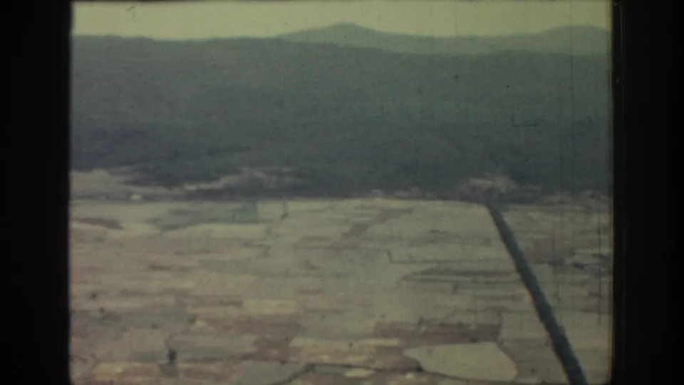 Rhodes 1962 home-movie still showing pale fields, a dark road or channel, and soft mountain silhouettes.