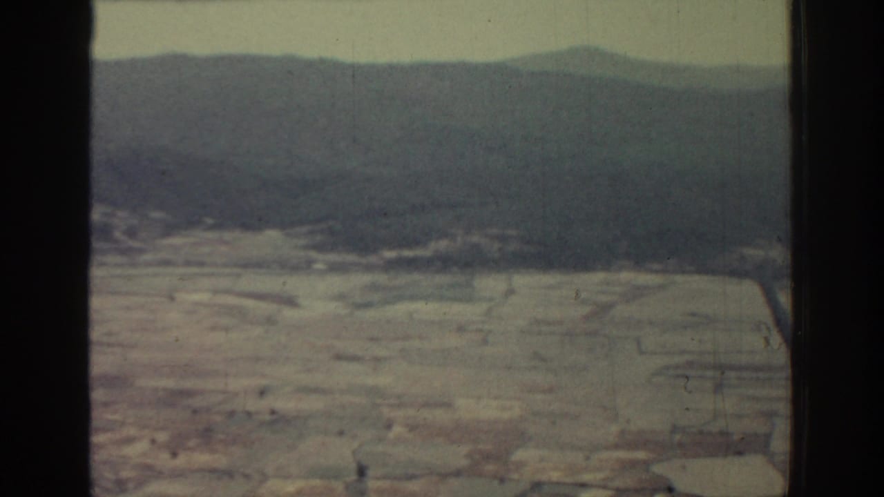 1962 Rhodes home-movie frame showing a hazy aerial view over pale fields, scattered tracks, and low dark hills in the distance.