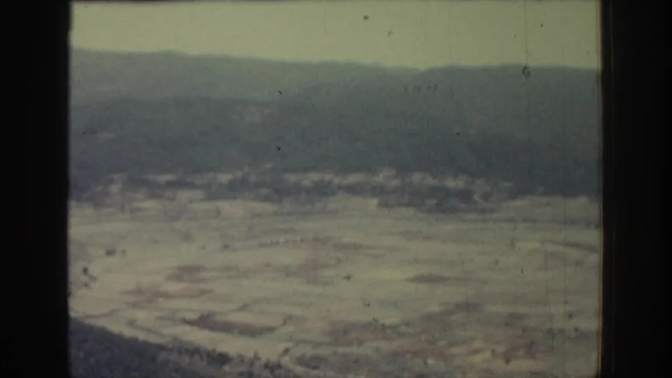 1962 Rhodes home-movie frame showing open rural fields, a dark lower edge, and hazy hills across the horizon.