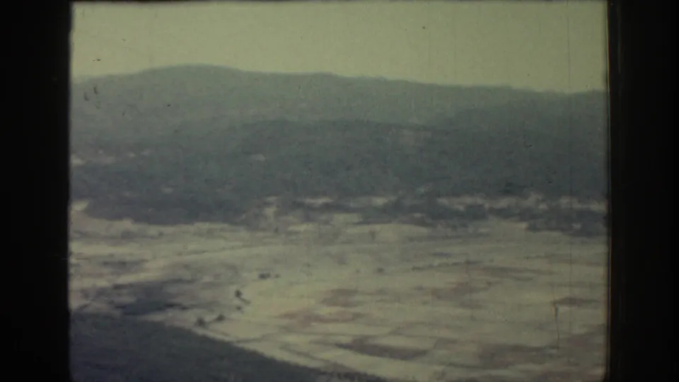 1962 Rhodes frame showing a pale patchwork of fields below hazy hills, with grain, scratches, and a dark lower frame edge.