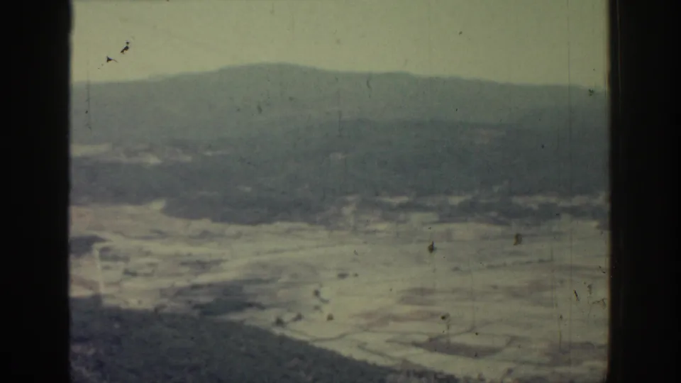 1962 Rhodes, Greece home-movie frame showing a hazy valley, wooded slopes and distant mountain ridges from an aerial viewpoint.