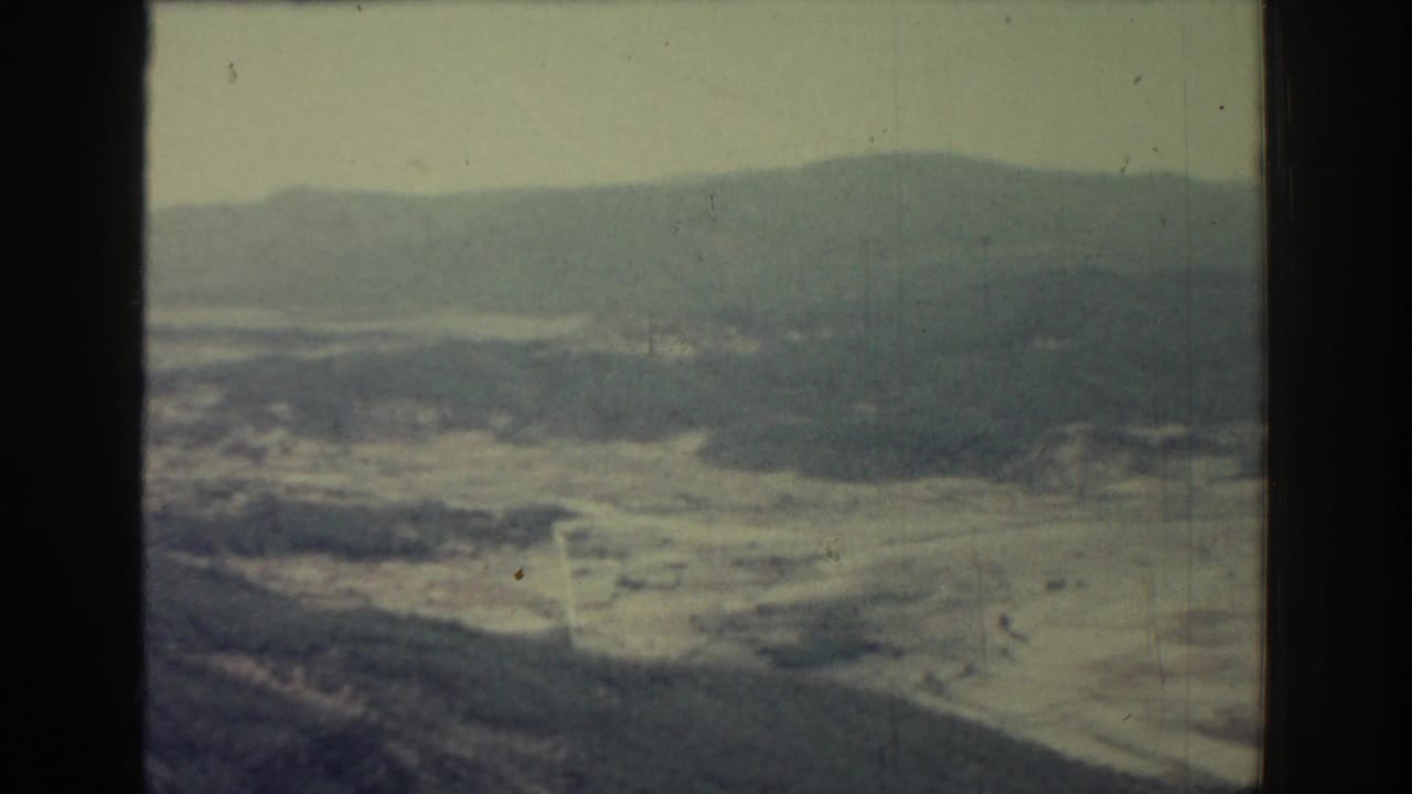 1962 Rhodes, Greece aerial home-movie view with rolling hills, a pale valley floor, scattered homes and soft mountain outlines.